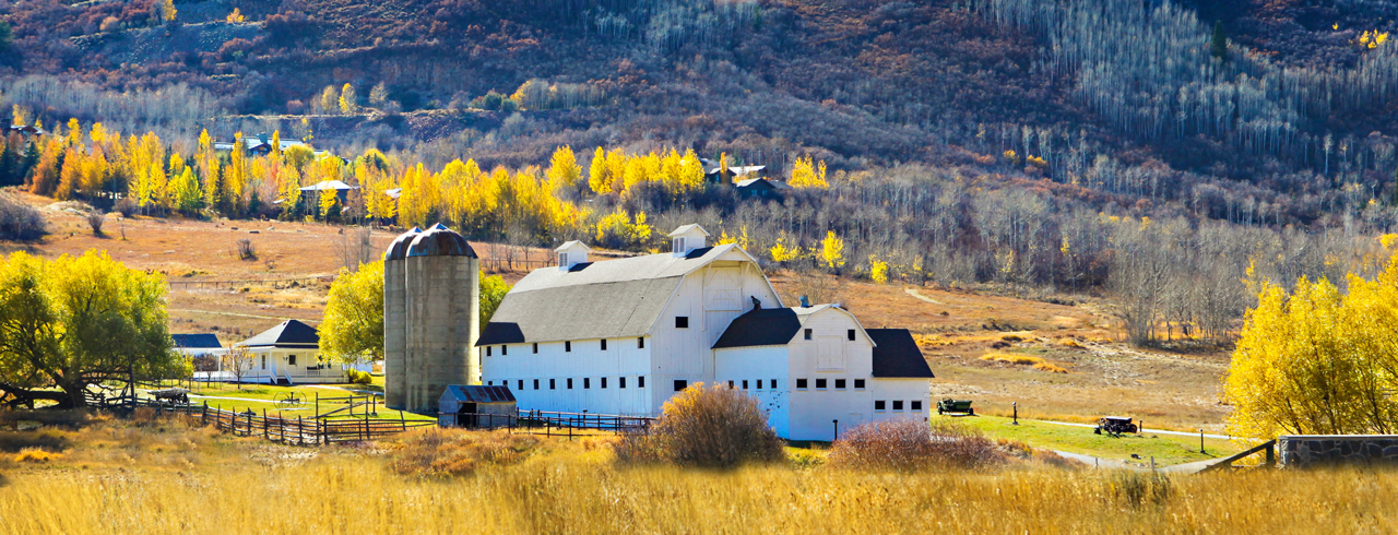 Rural Utah farm scene with a white barn, silo, golden fields, fall trees, and mountains in the background.