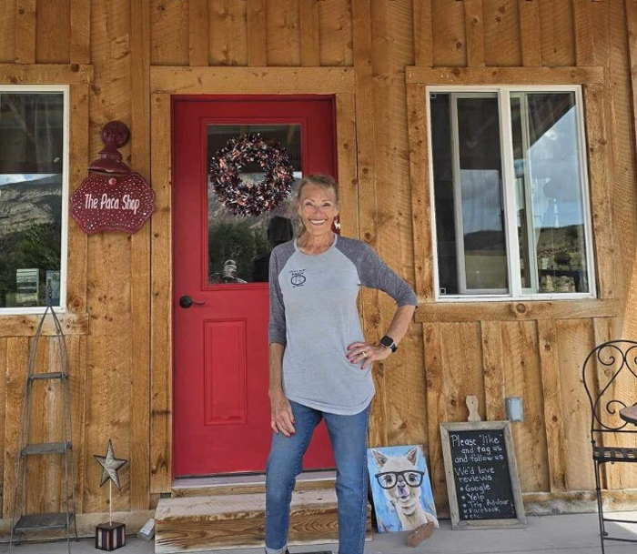 Smiling woman outside a rustic wooden building with a red door, alpaca artwork, a welcome sign and a sign for The Paca Shop.
