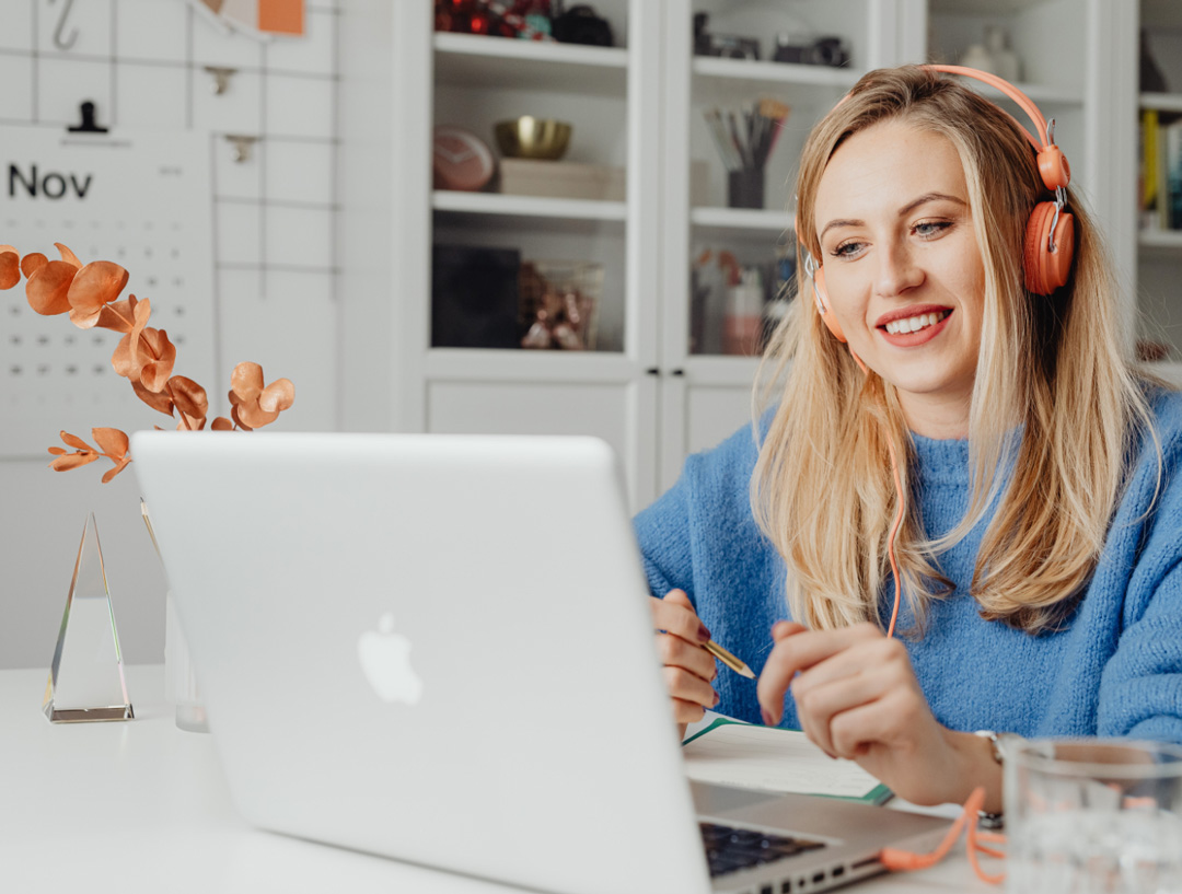 Smiling woman wearing orange headphones works from a laptop at her home office.