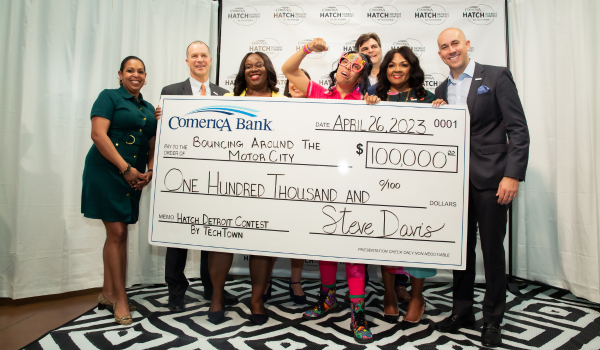 A group of people stand in front of a step-and-repeat with the Comerica Hatch Detroit Contest by TechTown logo on it. The group of people are smiling and holding a large check from Comerica Bank for $100,000, which is addressed to Detroit-based party rental and event planning business Bouncing Around the Motor City