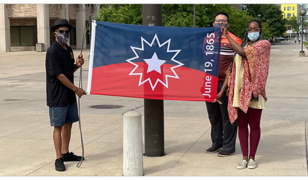 Three people wearing masks stand outside. Two people hold up the Juneteenth flag which is blue and red and features a large white star design in the center. The date June 19, 1865 is printed horizontally on the ride side of the flag