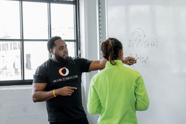 A person standing in front of a white board, pointing at a math equation with a dry erase market. A student stands in front of that person, looking at the equation