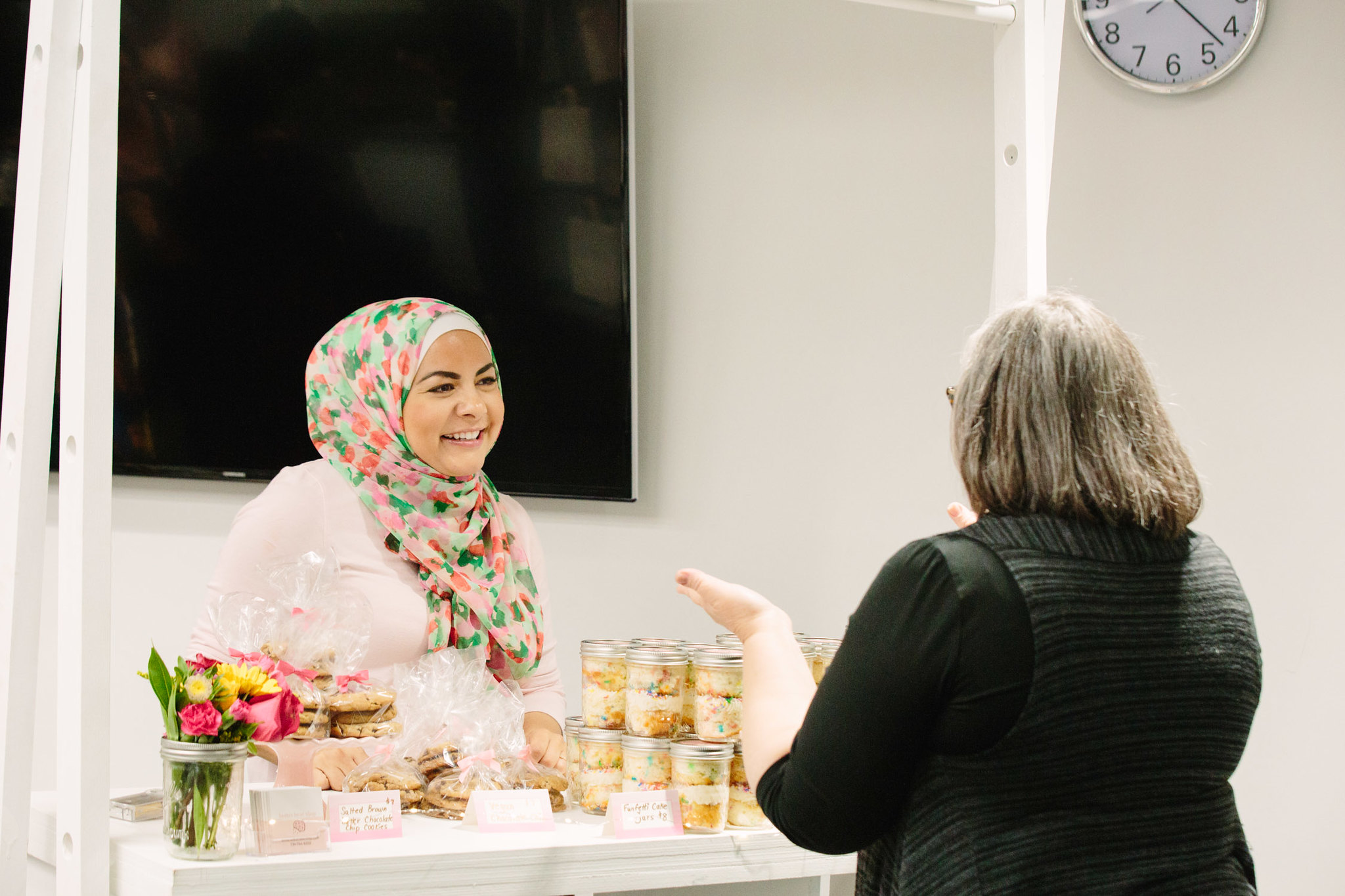 A woman speaks with another woman from behind her pop-up display of desserts