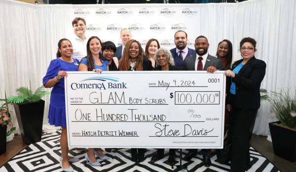 A group of people smile for a photograph, while a few of them hold a large check for $100,000 from Comerica Bank, which was awarded to G.L.A.M. Body Scrubs, the winner of the 2024 Comerica Hatch Detroit Contest by TechTown