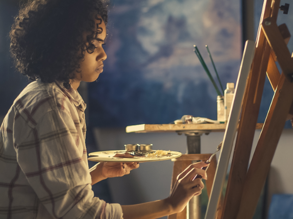 Person holding a palette and painting on a canvas propped on an easel