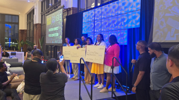 Six people standing on a raised platform, holding large checks